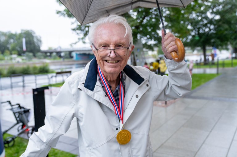 Eldre mann smiler til kamera mens han holder en paraply og har en medalje rund halsen.
