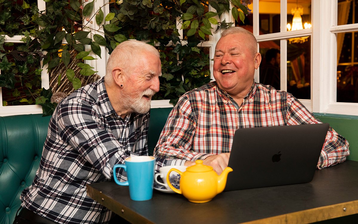 To eldre menn sitter på en kafe og smiler og ler. 