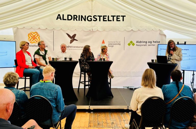 Fem personer sitter på høye stoler på en scene mens en sjette snakker i mikrofon