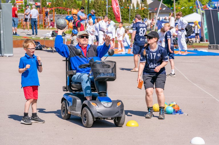 Eldre mann i en elekstrisk rullestolbil kaster håndball mens en ung dame og en liten gutt står ved siden av og heier