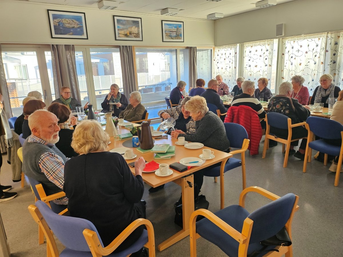 Flere personer sitter i et rom samlet i grupper rundt et bord. 