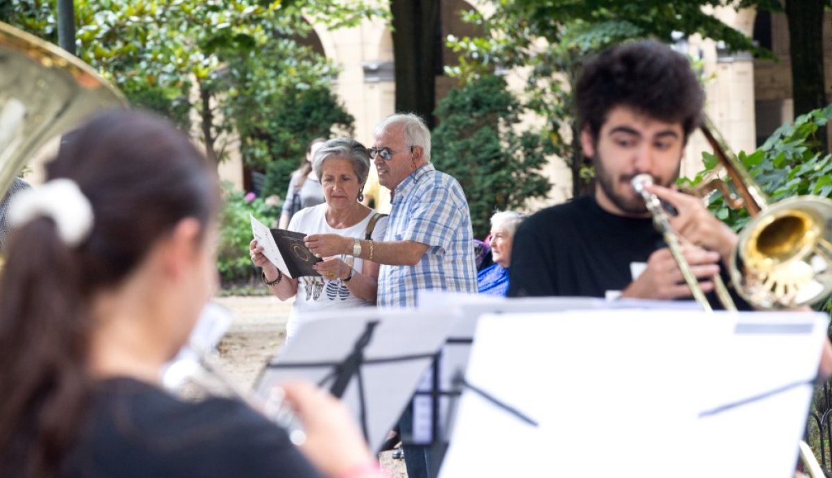 Yngre personer spiller musikk i forgrunnen mens det står et eldre par litt lengre bak i bildet og ser på en brosjyre sammen