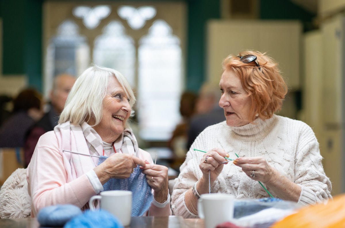 To eldre damer sitter å strikker, ser på hverandre og smiler. 