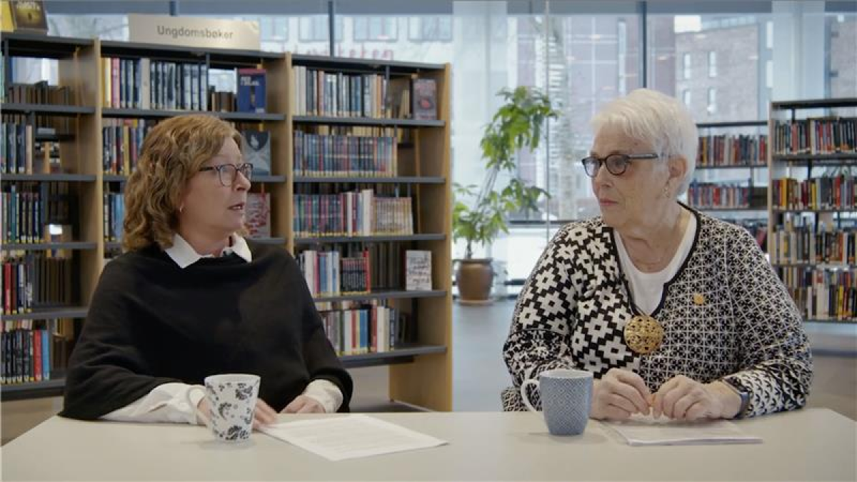 Trude og Kari sitter på hver sin stol ved et bord i et bibliotek.