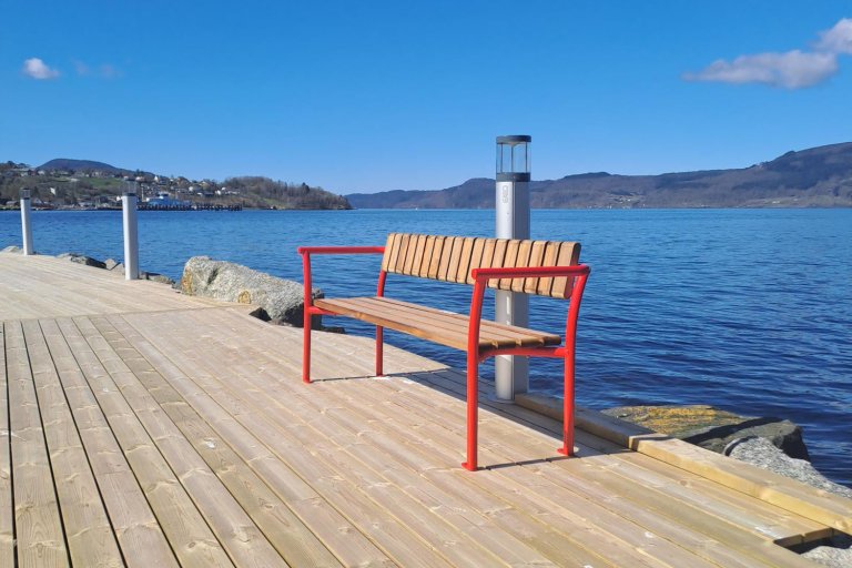 Benk med røde armlener plasser på en bryggekant med sjøen i bakgrunnen