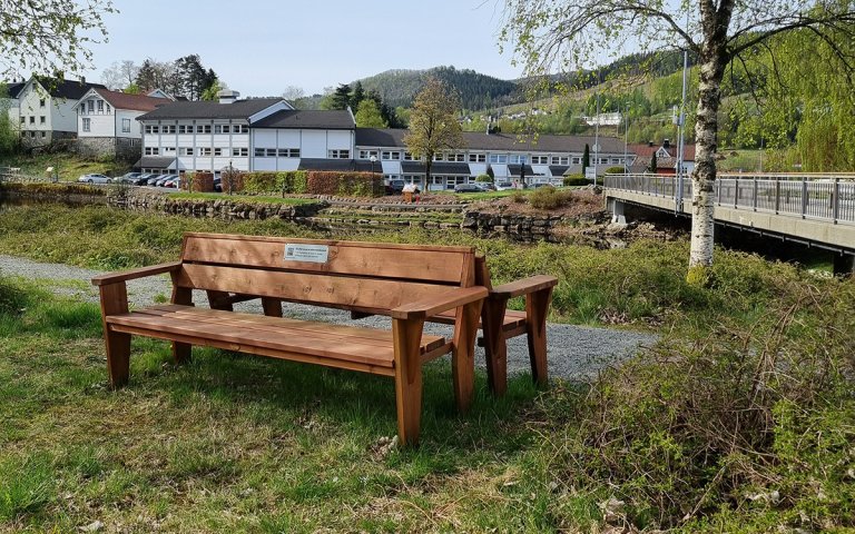 To aldersvennlige benker står rygg mot rygg. Den ene ut mot en grussti som følger en elv, den andre ut mot gresset.