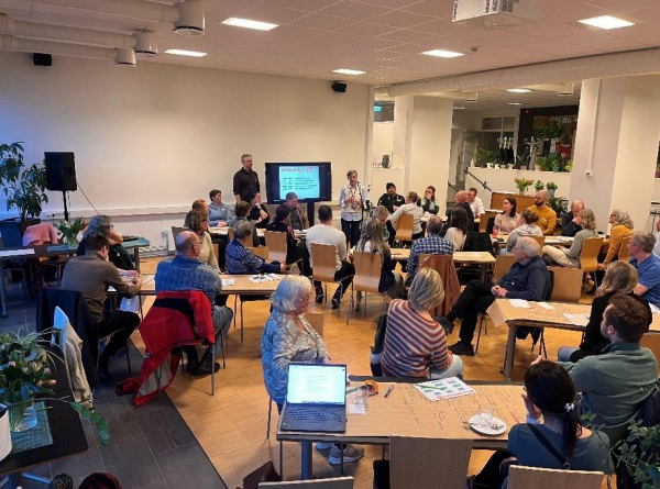 Et møterom der en står og presenterer og flere mennesker sitter i grupper rundt bord. 