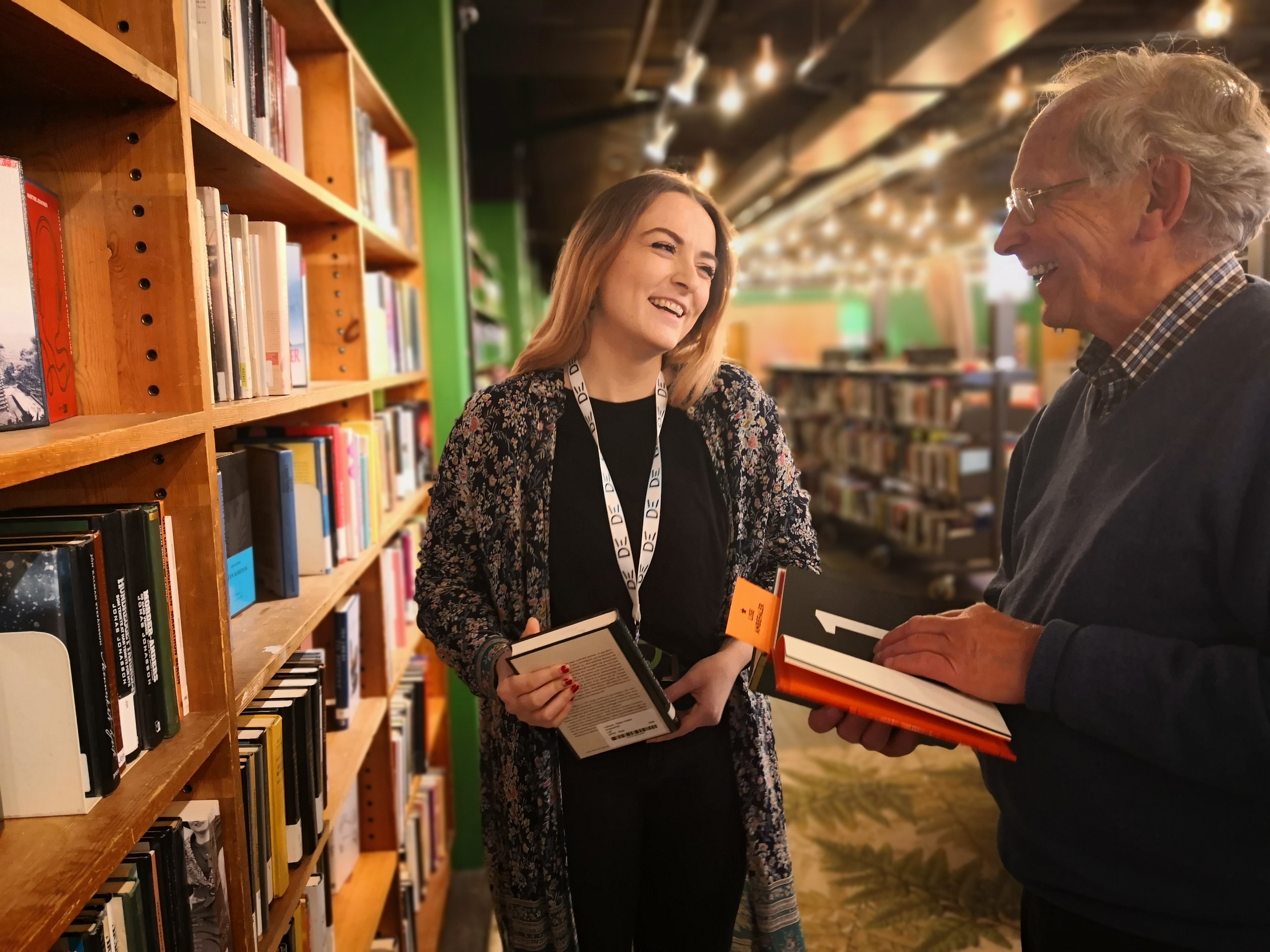 En dame og eldre mann står ved en hylle i biblioteket og smiler til hverandre med bøker i hendende