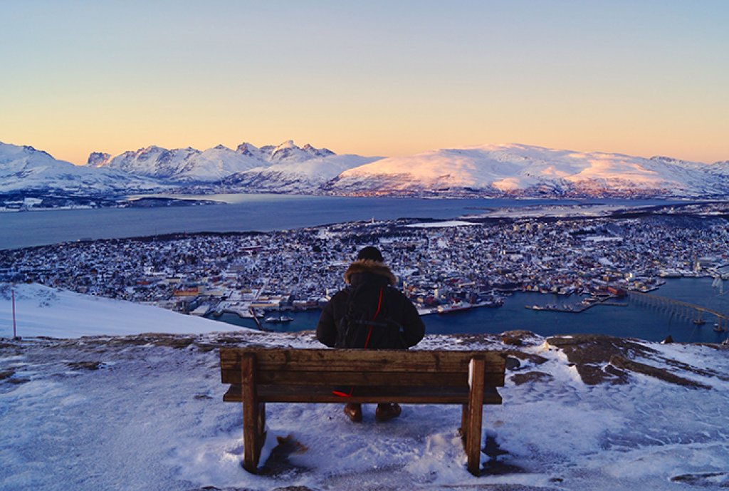 Mann sitter på en benk på toppen av et fjell en sein ettermiddag og ser utover en snødekt by. 