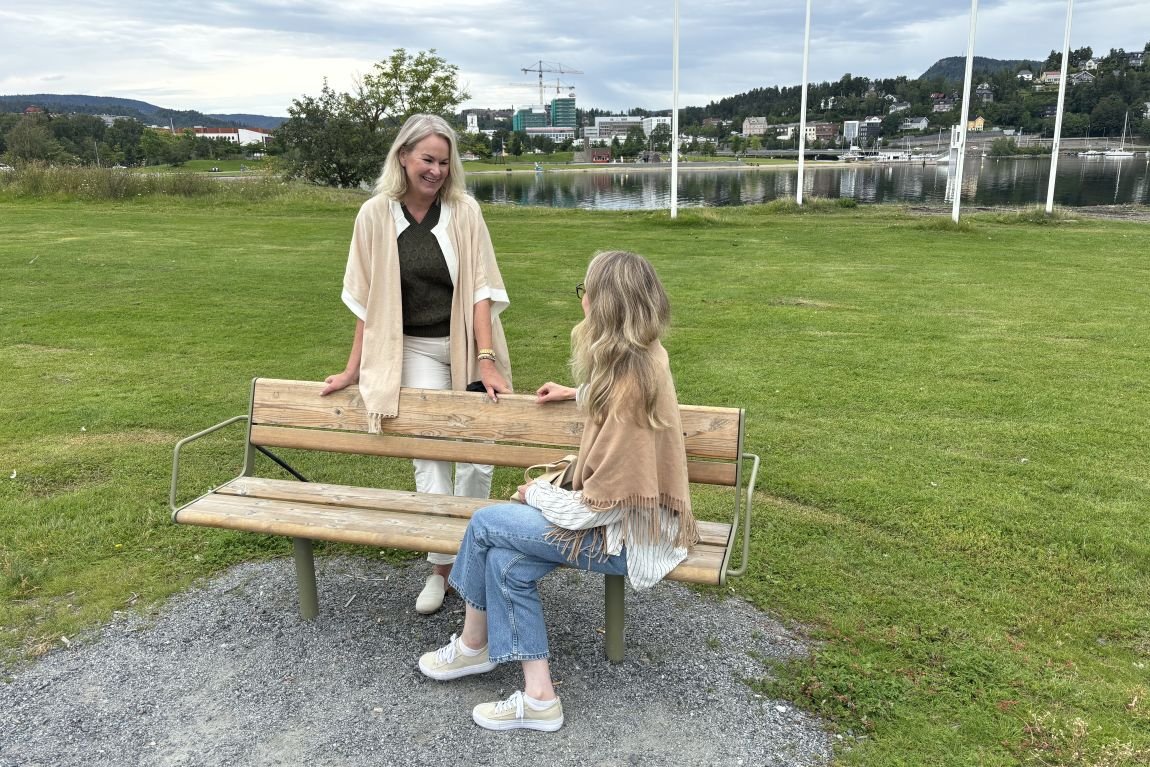 En dame står bak en benk og en dame sitter på en benk utendørs. De snakker med hverandre. 