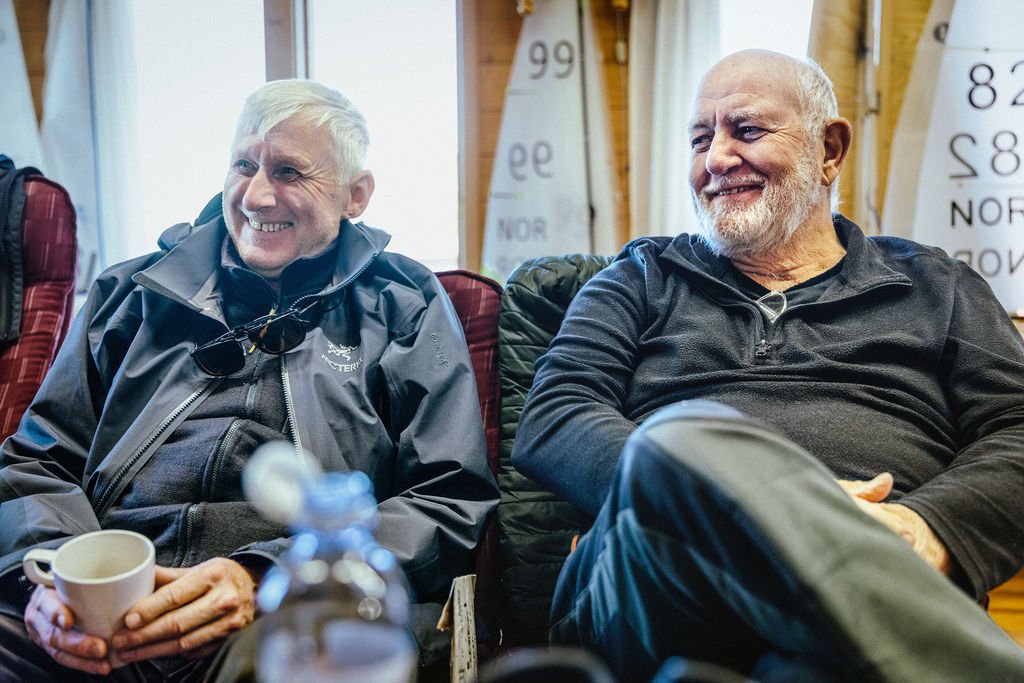 To eldre menn sitter i en sofa og drikker kaffe og smiler