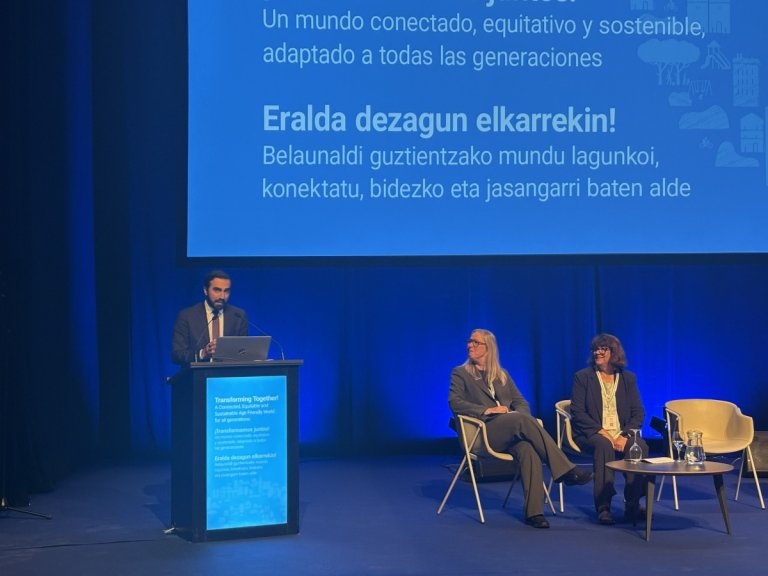 Usman står ved en talerstol på en stor scene der det sitter to personer