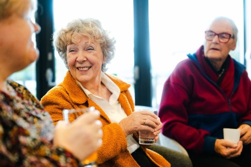tre eldre personer sitter ved siden av hverandre og smiler, holder hvert sitt glass eller kopp med drikke